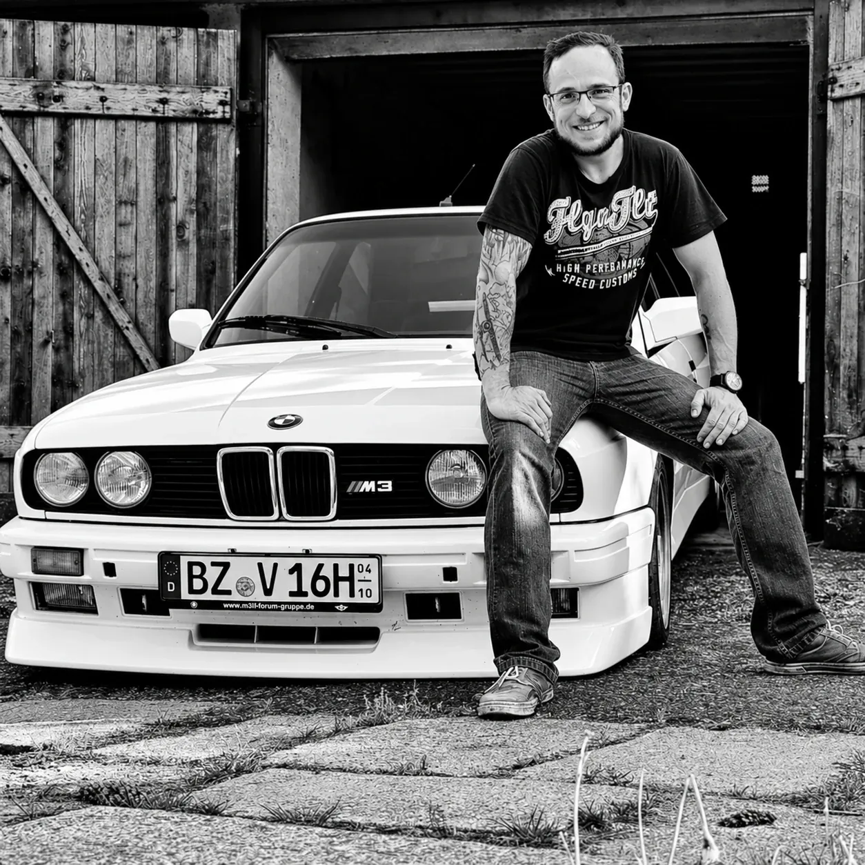 Patrick Pölchen, KÜS-Prüfingenieur und Inhaber von deinPRÜFER.de, auf der Motorhaube seines weißen BMW E30 M3 vor einer Holzgarage – Schwarz-Weiß-Foto